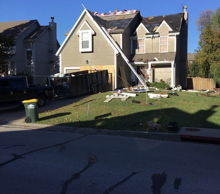 House with storm damage, missing roof shingles, construction debris in yard, and trailer parked nearby, illustrating the impact of Kansas City weather on roofing conditions.