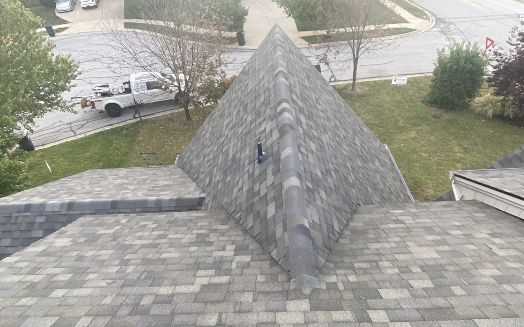 Roofing shingles on a residential home, showing a sloped roof structure with a vent, surrounded by trees and a street view, emphasizing home improvement and roof maintenance.